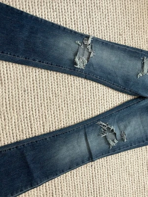 Blå slitna bootcut jeans - Säljer ett par blå jeans med slitningar och hål på framsidan. Klassisk femficksmodell med dragkedja och knapp. Jeansen har en tvättad finish och bootcut ben. Perfekta för dig som gillar streetstyle och trendiga detaljer.