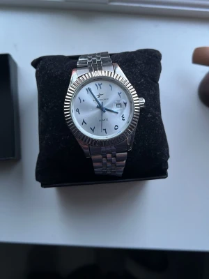 Silverfärgad klocka med arabiska siffror - Stilren klocka från Geneva i silverfärg med metallarmband och räfflad boett. Urtavlan är vit med arabiska siffror och datumvisning. Klassisk design som passar till många stilar. Fungerar felfritt