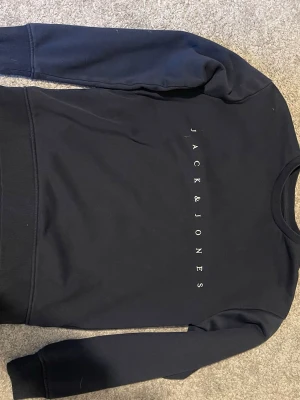 Blå sweatshirt från Jack & Jones - Snygg blå sweatshirt från Jack & Jones med vitt tryck på bröstet. Tröjan har rund halsringning, långa ärmar och ribbade muddar vid ärmslut och nederkant. Tillverkad i mjukt och bekvämt material, perfekt för en avslappnad stil.