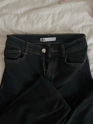 Svarta raka jeans från Zara - Säljer ett par svarta jeans från Zara med raka ben och kontrastsömmar. Klassisk femficksmodell med knappgylf och bälteshällor. Jeansen har en stilren look och passar perfekt till en trendig vardagsoutfit.