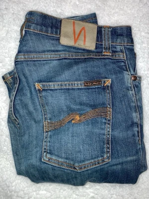 Blå jeans från Nudie Jeans - Säljer ett par klassiska blå jeans från Nudie Jeans med snygga kontrastsömmar och vågig brodyr på bakfickan. Jeansen är tillverkade i 100% ekologisk bomull och har en patch med logga bak i midjan. Perfekta för dig som gillar stilrena och tidlösa jeans.