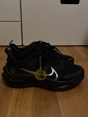 Nike ZoomX svarta sneakers helt nya - Snygga svarta Nike ZoomX sneakers med silverfärgade swoosh-detaljer och smiley-tag på sidan. Skorna har en chunky sula och mesh-överdel för extra komfort. Perfekta för dig som gillar sportig och modern stil.