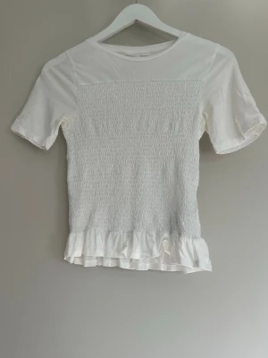 Vit topp med volang ifrån Gina Young - Säljer denna vita smockade topp med volang ifrån Gina Young i storlek 170.Toppen är använd ett antal gånger men har inga defekter.