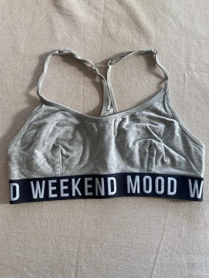 Grå sport-bh med textdetalj från ginatricot - Grå sport-bh i mjukt bomullsmaterial med svarta resårband där det står 'WEEKEND MOOD' i vita bokstäver. Bh:n har brottarrygg och bred resår under bysten för extra stöd. Perfekt för en avslappnad och sportig stil.
