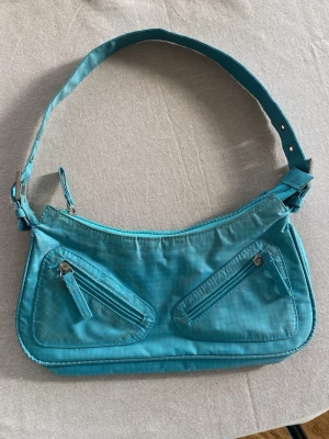 Turkos axelväska med dragkedjor från pieces - Snygg turkos axelväska i nylon med justerbart axelband och två sneda dragkedjefickor framtill. Väskan har en modern, avlång form och rutmönstrad yta som ger en cool retrovibe. Perfekt för dig som vill sticka ut med färg och smart design.