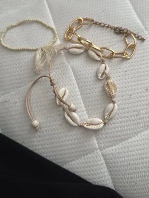 Set med armband i pärlor, snäckor och metall - Säljer ett set med tre armband: ett tunt pärlarmband i ljusgul ton, ett justerbart snäckarmband med beige snöre och vita snäckor samt ett chunky guldigt metallarmband med stora länkar. Perfekt för dig som gillar att mixa olika stilar och material.