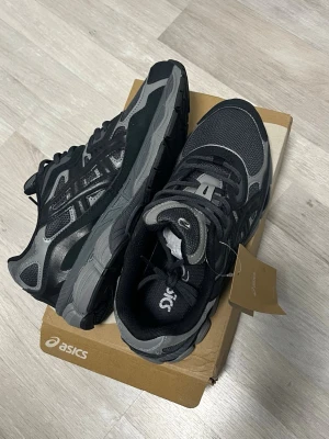 Svarta och grå sneakers från Asics - Snygga sneakers från Asics i svart och grå mesh och syntetmaterial. Skorna har klassisk låg profil, robust sula och coola detaljer på sidan. Perfekta för dig som vill ha en sportig och trendig look. Skorna har Asics-logga på tungan och sidan.