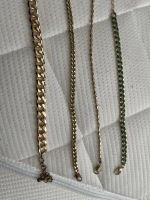 Fyra kedjearmband i guldton - Säljer fyra stilrena kedjearmband i guldton, ett av dem har inslag av grönt. Alla armband har klassiska länkar och metallfinish. Perfekta för att ge din outfit en extra touch av stil. Märke ej angivet.
