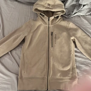 Beige huvtröja från Sail Racing - Snygg beige huvtröja från Sail Racing med dragkedja och fleecefoder. Perfekt för kyliga dagar med sin mysiga insida. Tröjan har en praktisk bröstficka med dragkedja och en justerbar huva.