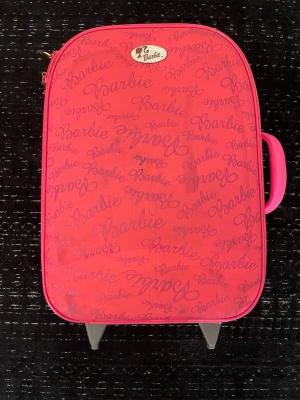 Rosa Barbie resväska med hjul - Säljer en rosa resväska från Barbie med hjul och draghandtag. Väskan har ett mönster med Barbie-loggan i olika storlekar över hela ytan och ett praktiskt bärhandtag på sidan. Perfekt för dig som vill resa med stil och älskar Barbie.