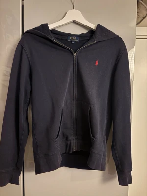 Marinblå hoodie från Polo Ralph Lauren - Snygg marinblå hoodie från Polo Ralph Lauren med dragkedja framtill och klassisk röd logga broderad på bröstet. Hoodien har huva och två fickor. Tillverkad i mjukt bomullsmaterial för skön känsla. Nackdelen är att en del på dragkedjan har lossnat.  (kolla bild 3) Passar också M skulle jag tro. 