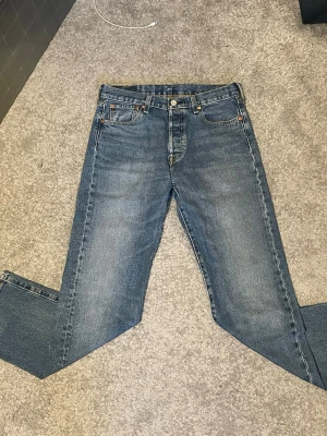 Levi's 501 blå jeans W30 L32 - Klassiska blå Levi's 501 jeans med rak passform och knappgylf. Jeansen har fem fickor och är tillverkade i slitstark denim. Perfekta för en avslappnad och tidlös stil. Färgen är mellanblå med lätt tvättade detaljer.
