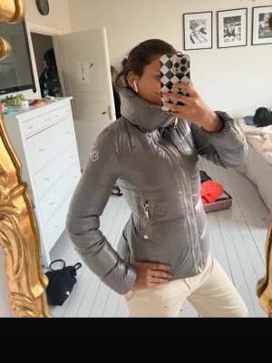 Grå dunjacka från Moncler - Snygg grå dunjacka från Moncler med hög krage och dragkedja framtill. Jackan har två fickor med guldfärgade dragkedjor och Moncler-logga på ärmen. qr kod finns inuti men inget annat äkthetsbevis där av pris 