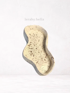 beige dekorbricka - Säljer en dekorbricka i organisk form. Brickan är vit med beige stänk och har en unik, böljande design. Perfekt som smyckesfat eller för småprylar. Tillverkad i lera.