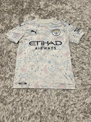 Manchester City paisley matchtröja - Säljer en Manchester City matchtröja från Puma med unikt paisleymönster i ljusblått och rosa på vit botten. Tröjan har korta ärmar, klubbmärke och sponsortryck framtill. Perfekt för dig som vill sticka ut på läktaren eller träningen.