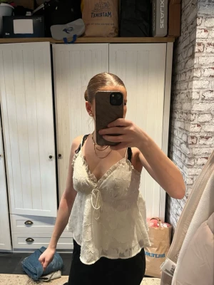 Vit spetslinnetopp med knyt - Säljer en vit linnetopp med spetsdetaljer och knyt framtill. Toppen har tunna axelband och v-ringning, vilket ger en feminin look. 