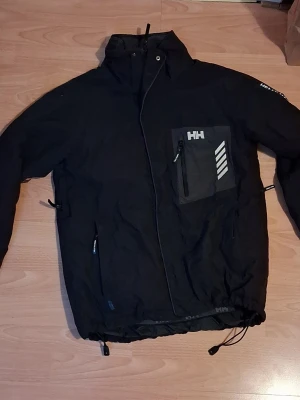 Helly Hansen Swift Jacket, en skidjacka och skidbyxor - Helly Hansen Swift Jacket, en skidjacka och skidbyxor  som kombinerar klassisk design med prestandamaterial. Den känns igen på den karaktäristiska bröstfickan med vita ränder och den vita HH-logotypen.  Evo  +1 Nyckelegenskaper: Material: Tillverkad i polyamid med Helly Tech-teknologi, vilket gör den både vind- och vattentät. Vattentäthet: Modellen har en vattenpelare på ca 14 000 mm. Funktioner: Ofta utrustad med Primaloft-isolering för värme, snölås, liftkortsficka 