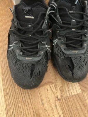 Svarta mesh sneakers med snörning - Säljer ett par svarta sneakers med ventilerande mesh-ovandel och snörning. Skorna har en sportig look med reflekterande detaljer och låg profil. Perfekta för dig som gillar streetwear och vill ha något bekvämt till vardags.