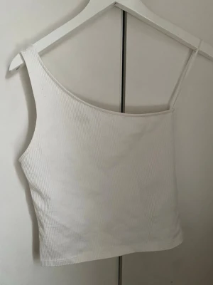 Vit oneshoulder ribbad topp - Säljer en vit ribbad topp med oneshoulder-design och smalt axelband på ena sidan. Toppen är croppad och har en clean, modern look som passar perfekt till jeans eller kjol. Materialet är mjukt och stretchigt för en bekväm passform.