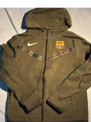 Olivgrön FC Barcelona hoodie Nike - Snygg olivgrön hoodie från Nike med FC Barcelona-logga på bröstet och dragkedja framtill. Tröjan har huva, långa ärmar och diskret mönster över bröstet. Perfekt för dig som älskar fotboll och vill ha en sportig stil.