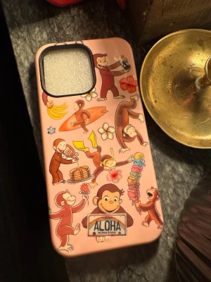 Rosa mobilskal iPhone 15pro Max  - Säljer ett rosa mobilskal dekorerat med flera illustrationer av Nyfikna Apan i olika roliga situationer, bland annat med glass, surfbräda och blommor. Skalets design har även en Hawaii-skylt och färgglada detaljer. Perfekt för dig som vill ha ett lekfullt och unikt skal.