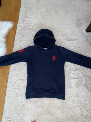 Mörkblå hoodie med röd logga - Snygg mörkblå hoodie från USPA med röd logga och text på bröstet samt en röd siffra på ärmen. Klassisk känguruficka framtill och huva. Perfekt för en avslappnad och sportig stil. Mjuk bomullskänsla och långa ärmar. Baksidan av tröjan har den jätte liten hål 