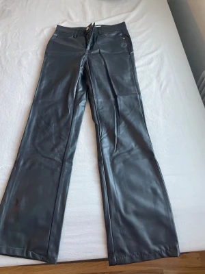 Svarta vida jeans i skinnlook - Säljer ett par svarta jeans med skinnlook och vida ben. De har klassisk femficksdesign, hög midja och knappstängning framtill. Materialet är glansigt och ger en cool känsla. Perfekt för dig som vill sticka ut med en trendig stil.