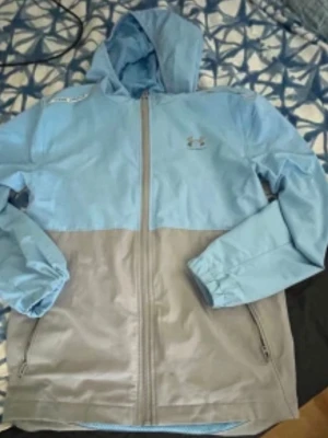 Blå och beige vindjacka från Under Armour - Snygg tvåfärgad vindjacka från Under Armour i ljusblått och beige. Jackan har huva, dragkedja framtill och två sidofickor med dragkedja. Perfekt för blåsiga dagar och enkel att matcha med olika outfits.