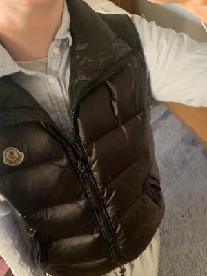 Moncler väst svart  - Snygg svart dunväst från Moncler med klassisk quiltad design och hög krage. Västen har två dragkedjefickor framtill och Moncler-logga på bröstet. Perfekt att slänga över en hoodie eller skjorta för en cool look.