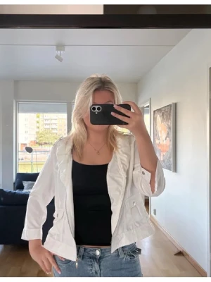 Vit jacka med detaljer  - Snygg vit jacka med dragkedja framtill och två fickor med lock och knapp. Jackan är kortare i modellen och har trekvartsärmar med knappdetalj vid ärmslutet. Den står som storlek 42 men är liten i storlek och passar mer som M/S