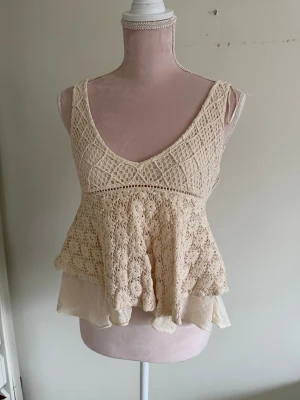 Beige spetslinne med volang - Supersött beige linne med spetsdetaljer och volangkant. Djup v-ringning både fram och bak, samt dubbla lager där det undre är i tunt tyg.  Köpt över 10 år sendan, finns inte kvar i sotetimentet och har aldrig sett någon med samma topp.