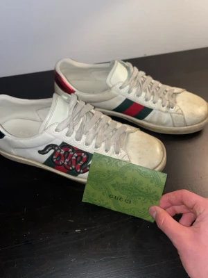 Gucci Ace sneakers med orm - Säljer ett par vita Gucci Ace sneakers med klassiska gröna och röda sidband samt en broderad röd orm på sidan. Skorna har vita snören och detaljer i metallicrött på hälen. Tillverkade i läder med gummisula och ikonisk Gucci-logo under sulan.