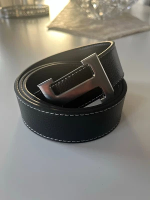 Svart skärp med H-spänne - Snyggt svart bälte med ett stort H-format metallspänne och vita kontrastsömmar längs kanterna. Bältet har en stilren och modern look, perfekt för att lyfta vilken outfit som helst. Tillverkat i skinn med en exklusiv känsla.