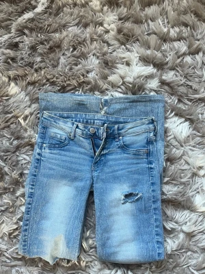 Ljusblå slitna bootcut jeans - Säljer ett par ljusblå bootcut jeans med slitningar och hål på benen för en trendig look. Jeansen har klassisk femficksmodell, låg midja och dragkedja med knapp. Perfekta för dig som gillar en avslappnad och edgy stil.