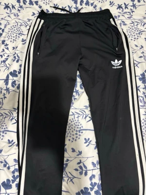 Svarta Adidas mjukisbyxor med ränder - Säljer ett par klassiska svarta mjukisbyxor från Adidas med vita ränder längs sidorna och logga på låret. Byxornaanen har snörning i midjan och dragkedjor vid fickorna. Perfekta för chill eller träning.