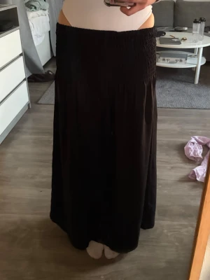 Svart långkjol med smock i midjan - Svart långkjol med bred smock i midjan som ger en bekväm och flexibel passform. Kjolen har en rak och flowy siluett, perfekt att styla med crop tops eller stickade tröjor. Enkel och stilren design utan mönster eller extra detaljer.