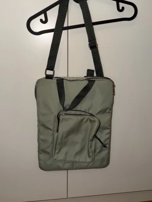 Grön datorväska med justerbar rem - Snygg datorväska i ljusgrönt med svart justerbar axelrem och praktiskt ytterfack med dragkedja. Väskan är rektangulär och har en enkel, stilren design som passar perfekt för att bära din laptop. Tillverkad i slitstarkt syntetmaterial.