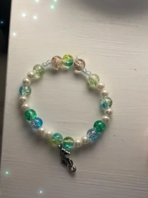 Färgglatt pärlarmband med sjöhästberlock - Säljer ett handgjort armband av mig med vita och färgglada pärlor i blått, grönt och gult. Armbandet har en silverfärgad sjöhästberlock. Omkrets 18 cm❤️