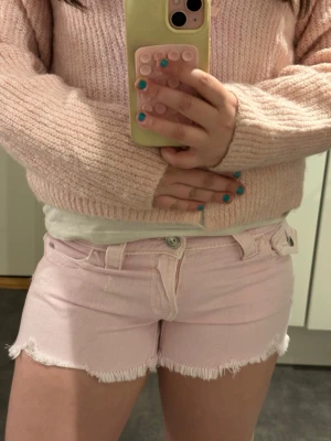 Ljusrosa jeansshorts med fransig kant - Säljer ett par ljusrosa jeansshorts med fransig kant och klassisk femficksdesign. Shortsen har bälteshällor och knappgylf. Perfekta för varma dagar och ger en söt och avslappnad look.