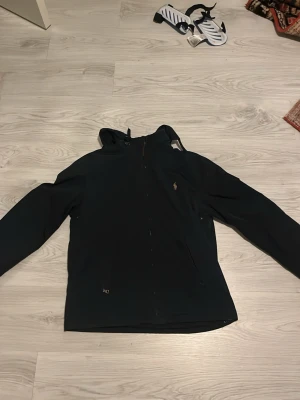 Svart vindjacka från Polo Ralph Lauren - Snygg svart vindjacka från Polo Ralph Lauren med huva och dragkedja framtill. Jackan har den klassiska Polo-loggan broderad på bröstet och två sidofickor med dragkedja. Tillverkad i ett lätt och vindtätt material, perfekt för blåsiga dagar.