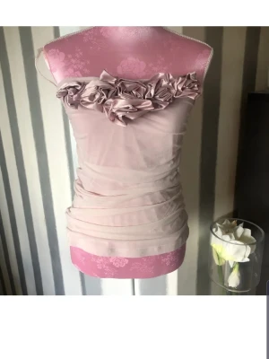 Ljusrosa topp med volangdetaljer - Supersöt ljusrosa topp med axelbandslös design och stora volangdetaljer längs överkanten. Tunn, stretchig mesh som ger en figurnära passform och snygg rynkning i tyget. Perfekt för dig som vill sticka ut lite extra.