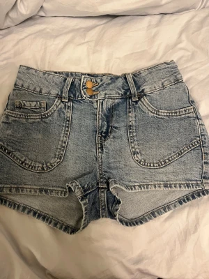 Blå denimshorts med broderade fickor - Säljer ett par blå denimshorts som har snygga broderade hjärtan på bakfickorna. Perfekta för sommaren och ger en avslappnad men trendig look. De är justerbara i midjan. Säljer likadana i grå i annan storlek!
