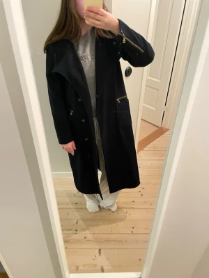 Svart lång ullkappa med guldknappar - Stilren svart lång ullkappa med stora guldknappar vid halsen och dragkedjor i guld på ärmar och fickor. Kappan har en rak passform, stora fickor framtill och hög krage som kan bäras både öppen och stängd. Perfekt för dig som gillar en klassisk men edgy look. Mycket fint skick men ett litet hål på insidan som lätt fixas med nål och tråd(se bild).