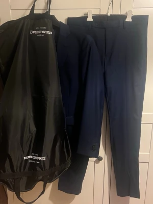 Mörkblå kostym från Dressmann - Snygg mörkblå kostym från Dressmann med klassisk kavaj och matchande byxor. Kavajen har två knappar, smala slag och fickor med lock. Perfekt för dig som vill ha en stilren och tidlös look. Kostymen Slim Fit 48, byxorna 32/32. Kostymen skickas bra förpackat och även i kostympåsen från Dressmann
