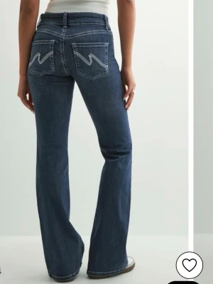 Nelly jeans - Mörkblå bootcut jeans med vita broderade detaljer på bakfickorna. Klassisk femficksmodell med hög midja och dubbla knappar framtill. Tillverkade i stretchig denim för skön passform. Lite slitna vid hälen men annars bra skick