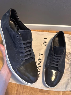 Svarta Lanvin sneakers med lackdetalj - Snygga svarta sneakers från Lanvin med klassisk låg profil. Skorna har ovandel i mocka och tåparti i blankt lackläder, samt vita gummisulor. Svarta skosnören och diskret design gör dem stilrena och lätta att matcha till olika outfits. Super skick utöver en liten fläck på vänstra toeboxen, kolla närmare på bild 1 och 2. Dustbags ingår ej!! Uk size 15, så de stora, motsvarar typ 49/50hör av dig vid intresse!