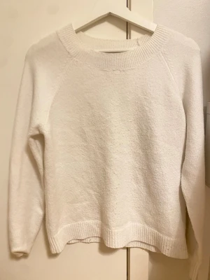 Vit stickad tröja med rund hals - En enkel och stilren vit stickad tröja med rund hals och långa ärmar. Tröjan har ribbstickade muddar vid hals, ärmslut och nederkant. Perfekt för lager-på-lager och passar till många olika outfits.