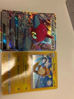 Pokémonkort Porygon-Z GX & Alolan Raichu - Två Pokémonkort: ett holografiskt Porygon-Z GX med färgstark bakgrund och ett Alolan Raichu med gul ram och glittrande effekt. Båda korten har tydliga illustrationer och detaljerade attacker. Perfekt för samlare eller Pokémon-fans. Det gula är Hollow. 