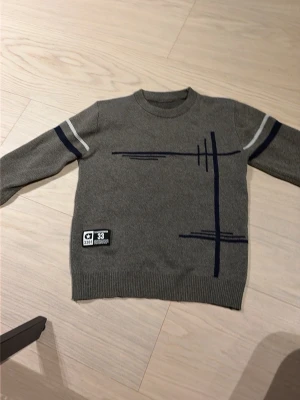 Grå stickad tröja med blå detaljer - Grå stickad tröja med rund hals och långa ärmar. Tröjan har mörkblå och vita ränder på ärmarna samt grafiska blå linjer framtill. Nedtill finns en liten patch med text. Perfekt för en stilren och avslappnad look.