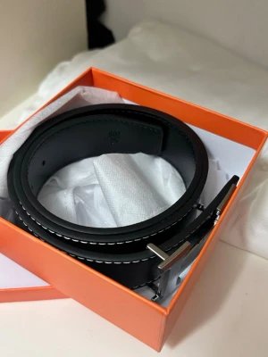 Svart skärp med silverfärgat spänne - Stilrent svart skärp i skinn med silverfärgat metallspänne och vita kontrastsömmar. Perfekt accessoar för att lyfta din outfit med en klassisk touch. Levereras i en orange presentask.